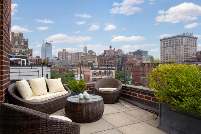 Rooftop terrace with wicker furniture, plants, and city skyline view under a blue sky with clouds.