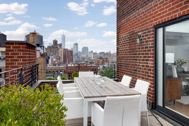Rooftop terrace with a wooden table and white chairs, overlooking city skyline with blue sky and clouds.