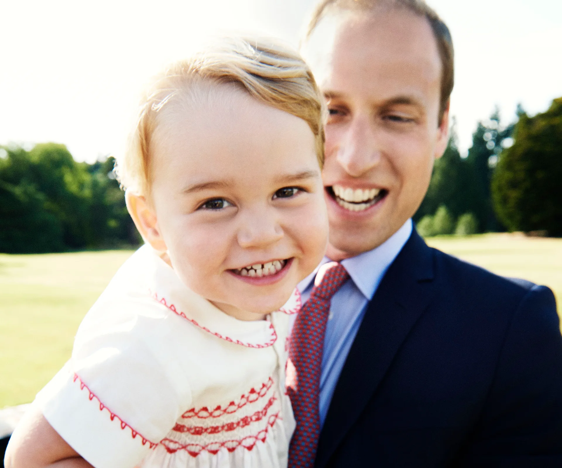 A child smiles while being held by an adult in a suit, outdoors in a sunny setting.