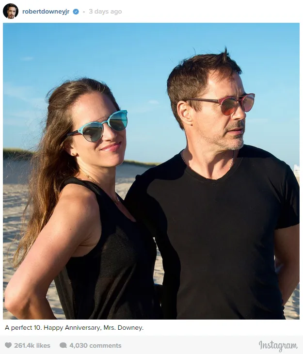 Man and woman in sunglasses standing on a beach, both wearing black shirts.