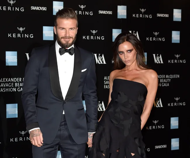 Man in a tuxedo and woman in a black strapless dress posing on a gala red carpet with a branded backdrop.