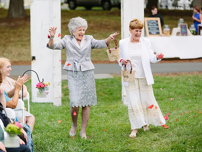 Two joyful older women throwing flower petals at an outdoor event with an audience.