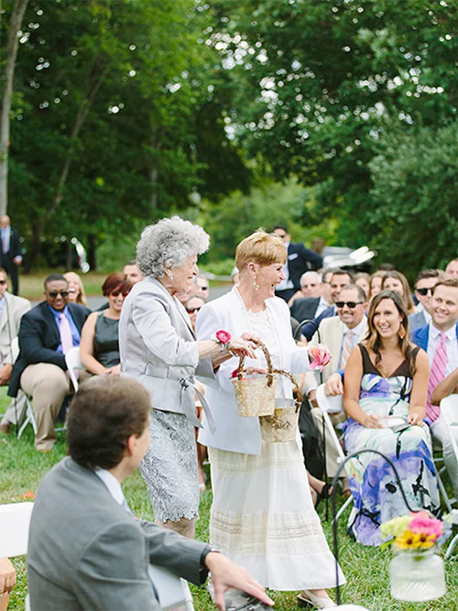 Two elderly women in light-colored outfits walk down an outdoor aisle, holding baskets, surrounded by seated guests.