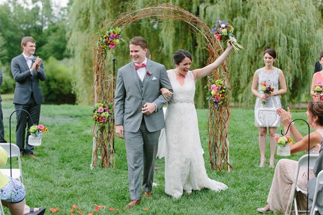 Bride and groom joyfully walk down the aisle under floral arch at outdoor wedding ceremony.