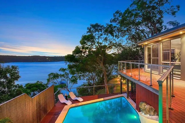 Hilltop house with pool overlooks a serene lake and lush trees at sunset.