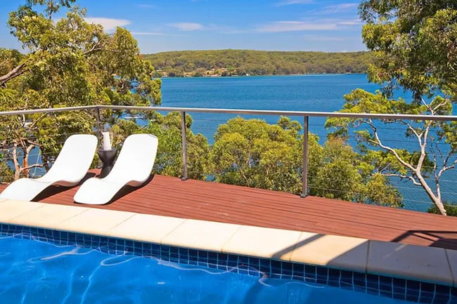Two white lounge chairs on a deck overlooking a pool, with a scenic view of a lake and forested hills in the background.