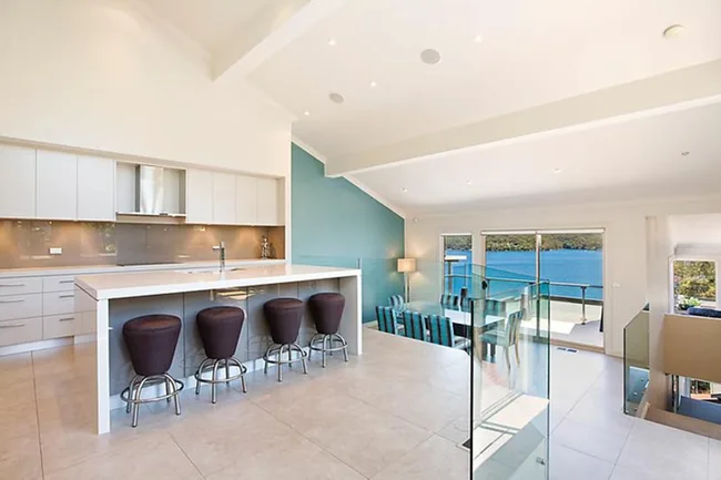 Modern kitchen with white cabinets, island, brown stools, dining area, large windows, and water view in the background.
