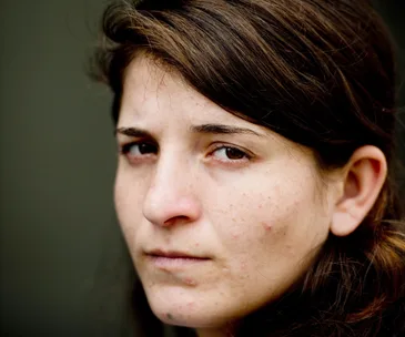 A woman with a somber expression, looking into the distance, close-up portrait with a plain background.