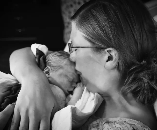 A woman holds a newborn baby close, both wrapped in a blanket, in a tender black and white photograph.