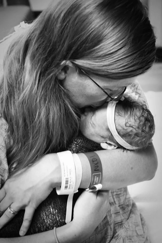 Mother embracing newborn baby, both wearing hospital wristbands, in a tender black and white moment.