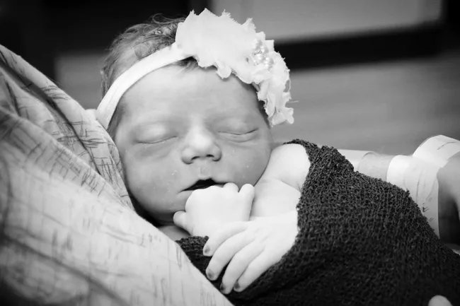 Newborn baby wrapped in a blanket, wearing a floral headband, peacefully sleeping in black and white.