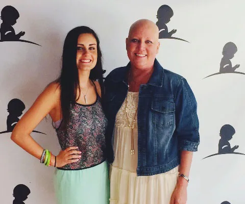 Two smiling women standing together, one with long hair and one bald, in front of a wall with child silhouettes.