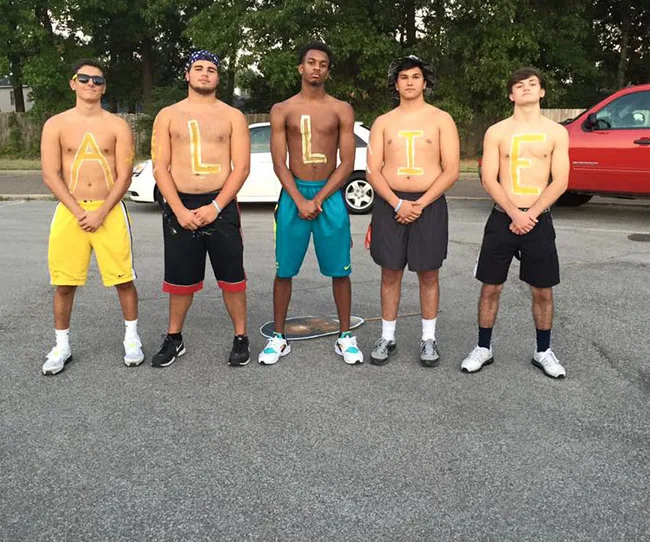 Five men stand shirtless with letters painted on chests, spelling "ALLIE," in a parking lot with trees and a red vehicle.