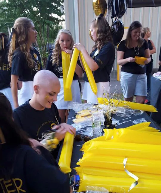 Young woman with shaved head smiling, surrounded by others, inflating yellow balloons at an event with decorations.