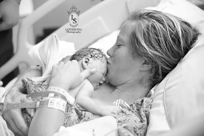 Mother lovingly holds and kisses her newborn baby in a hospital bed, captured in black and white photography.