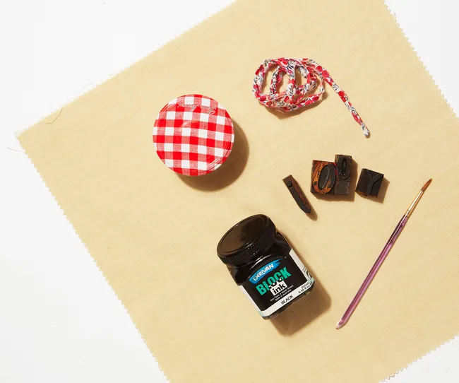 Art tools on fabric: red lid, patterned string, block ink jar, stamps, brush on beige fabric.