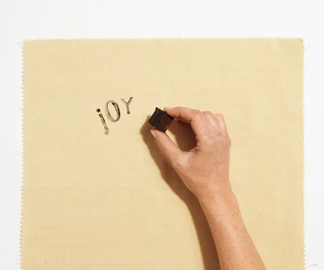 Hand stamping the word "joy" on beige fabric.