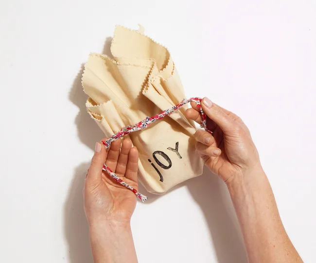 Hands holding a beige cloth bag with "joy" written on it, tied with a colorful floral string.
