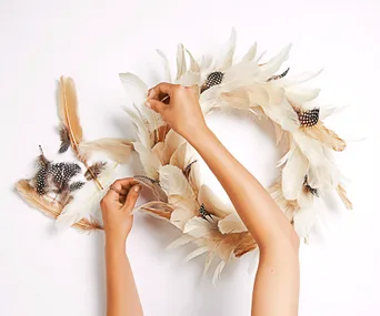 Hands crafting a feather wreath with white and brown spotted feathers on a white background.