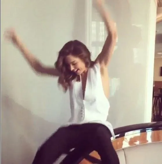 Woman in a white sleeveless top and black pants with arms raised, appearing joyful and seated indoors.
