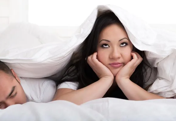 Woman looking bored under a blanket with sleeping man beside her.