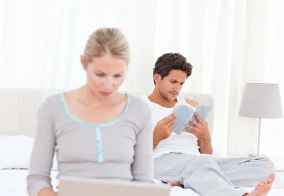 Man in white shirt reads a book on bed; woman in gray top uses a laptop. Background is bright and minimalistic.