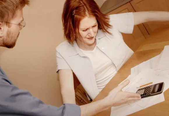 A woman and a man sit at a table with scattered bills and a calculator, both appearing stressed or concerned.