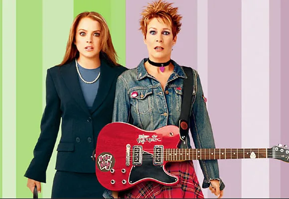 Two women stand together; one in a suit holding a briefcase, the other in a denim jacket with a red electric guitar.