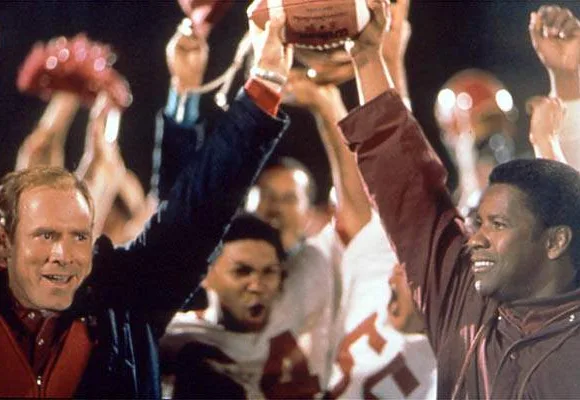 Two coaches celebrating with a football, surrounded by cheering football players.
