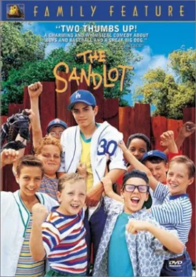 Group of boys in baseball attire stand together, smiling, in front of a fence with "The Sandlot" title overhead.