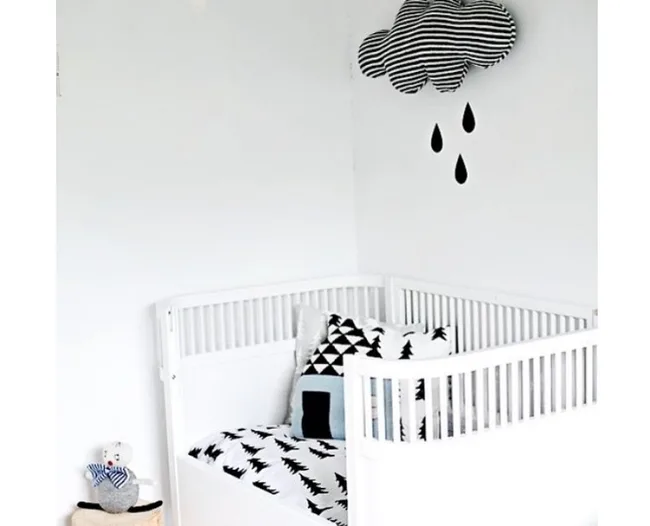 White baby crib with black and white bedding, cloud pillow above, and stuffed toy in corner.
