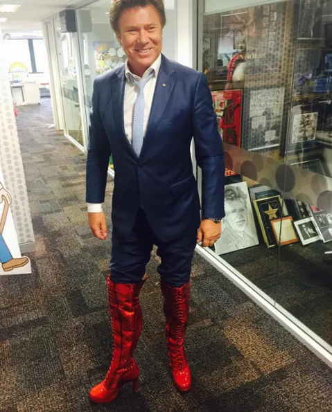 Man in blue suit and red knee-high boots stands smiling in an office hallway.