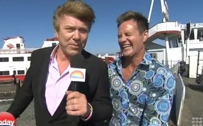 Two men laughing during an outdoor TV interview near a docked ferry on a sunny day.