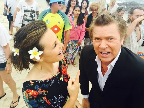 Two people smiling on a beach, woman with flowers in hair, surrounded by a crowd, ocean in the background.