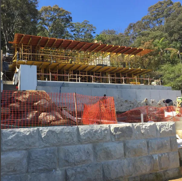 Construction site with scaffolding and orange safety netting on a hillside, with trees in the background.