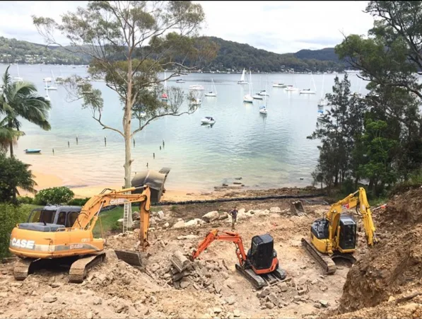 Excavators on a construction site near a scenic bay with sailboats and trees in the background.
