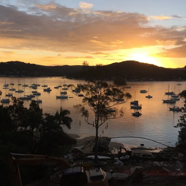 Sunset over a calm bay with numerous sailboats, silhouetted trees, and hills in the background.
