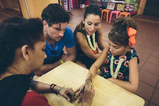 A girl getting henna applied, watched by a man and woman, in a colorful, indoor market setting.