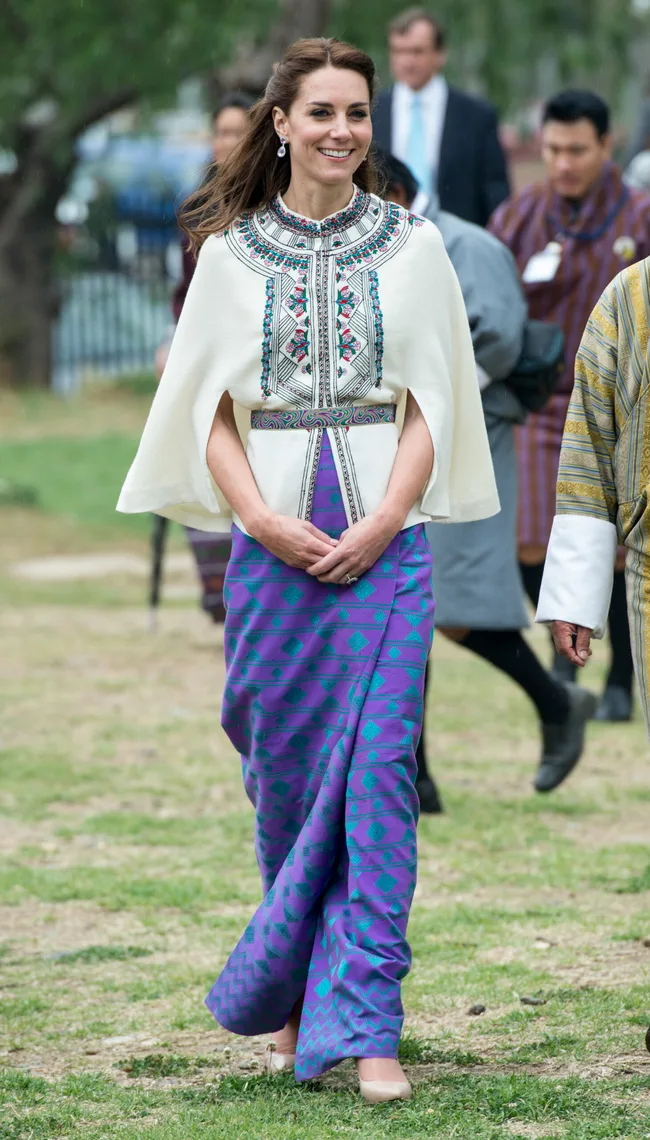 A woman in a colorful, traditional Bhutanese-inspired dress, smiling outdoors with people in the background.