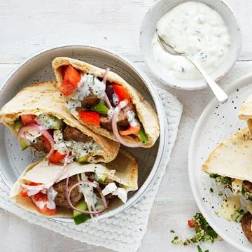 Pita wraps filled with meat, tomatoes, cucumbers, onions, and tzatziki sauce, served with a side of extra sauce.