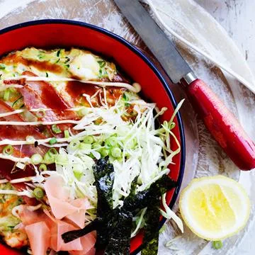 Red bowl with okonomiyaki topped with shredded cabbage, seaweed, pickled ginger, green onions, knife, and lemon wedge.