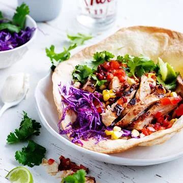 Grilled chicken taco in a tortilla bowl with red cabbage, corn, tomatoes, cilantro, and lime on a white plate.