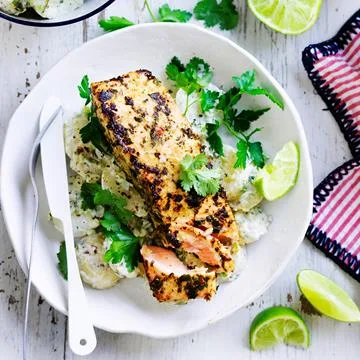 Grilled salmon on creamy potato salad, garnished with fresh herbs and lime wedges, served in a white bowl.