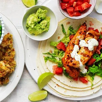 Grilled chicken tacos with lettuce, diced tomatoes, and sauce, served with guacamole, lime wedges, and a side dish.