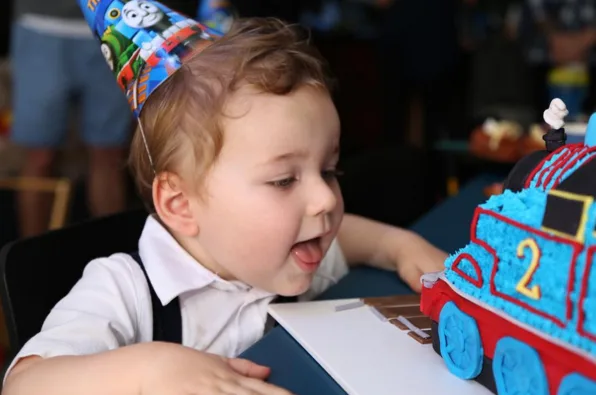 Child in birthday hat excitedly looking at a Thomas the Tank Engine-themed cake.