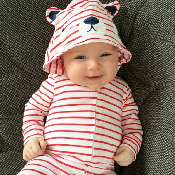 Baby in a red and white striped outfit with a hood featuring bear ears and face, smiling on a dark cushion chair.