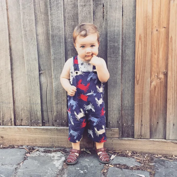Toddler in crab-patterned overalls stands in front of a wooden fence, with hand near mouth, wearing brown sandals.