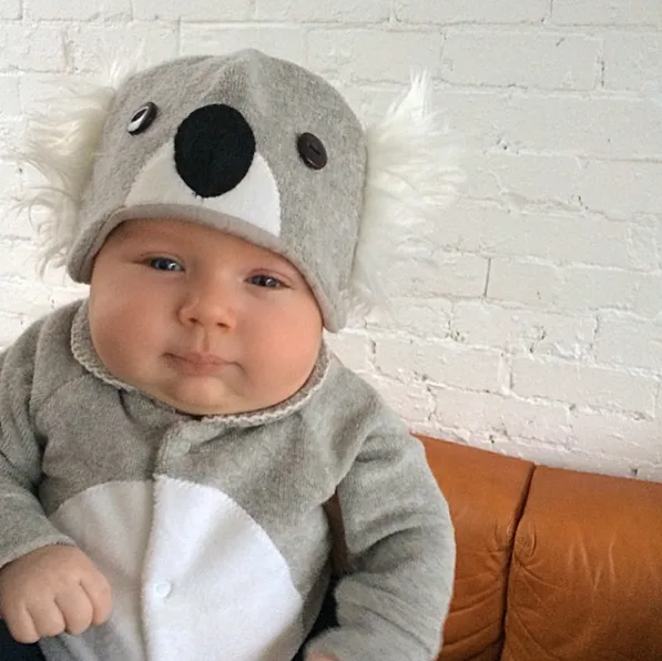 A baby wearing a grey koala outfit with a hat sits on a brown couch against a white brick wall.