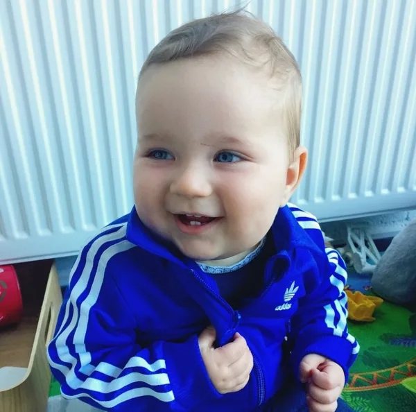 A baby smiling, wearing a blue Adidas tracksuit, sitting indoors.