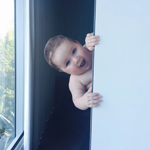 A joyful baby peeking from behind a white wall with a bright expression, near a window.